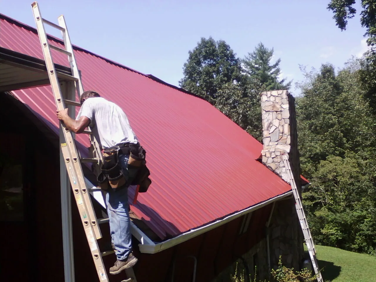 Roofer on a ladder beside a red metal roof and stone chimney — Soffit & Fascia Repair by Roofing Brothers in Andover